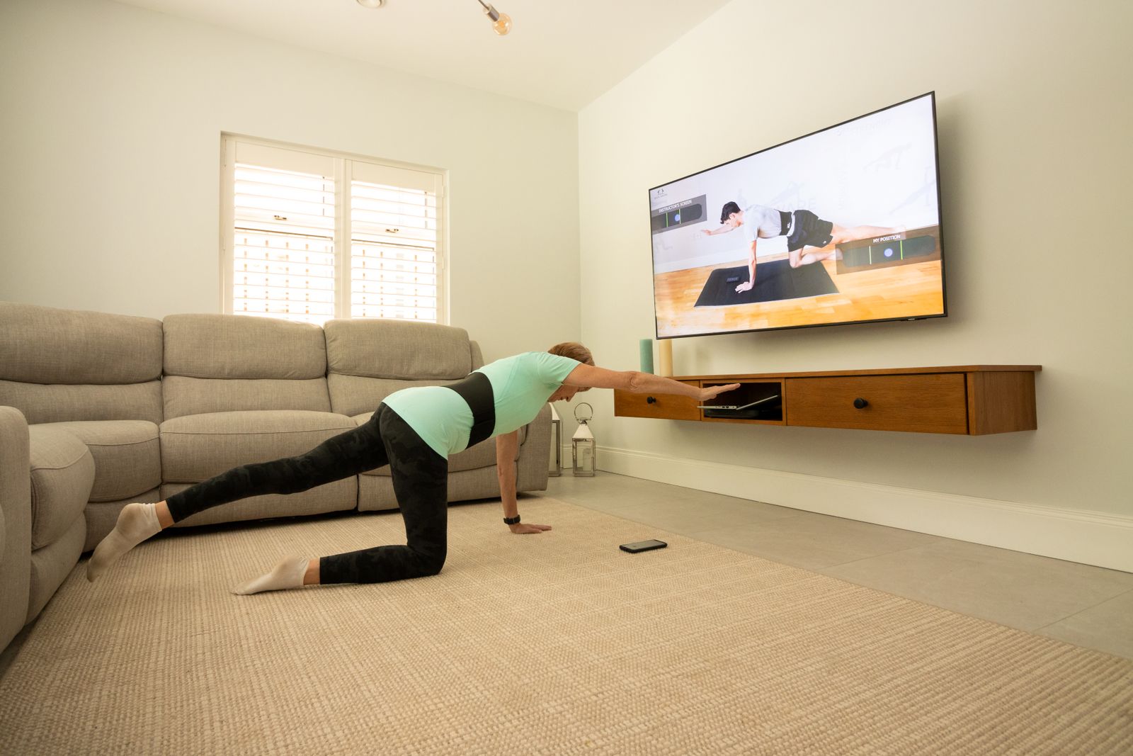 A woman holding a plank with one leg lifted, on a mat in a living room, following a class on the television.